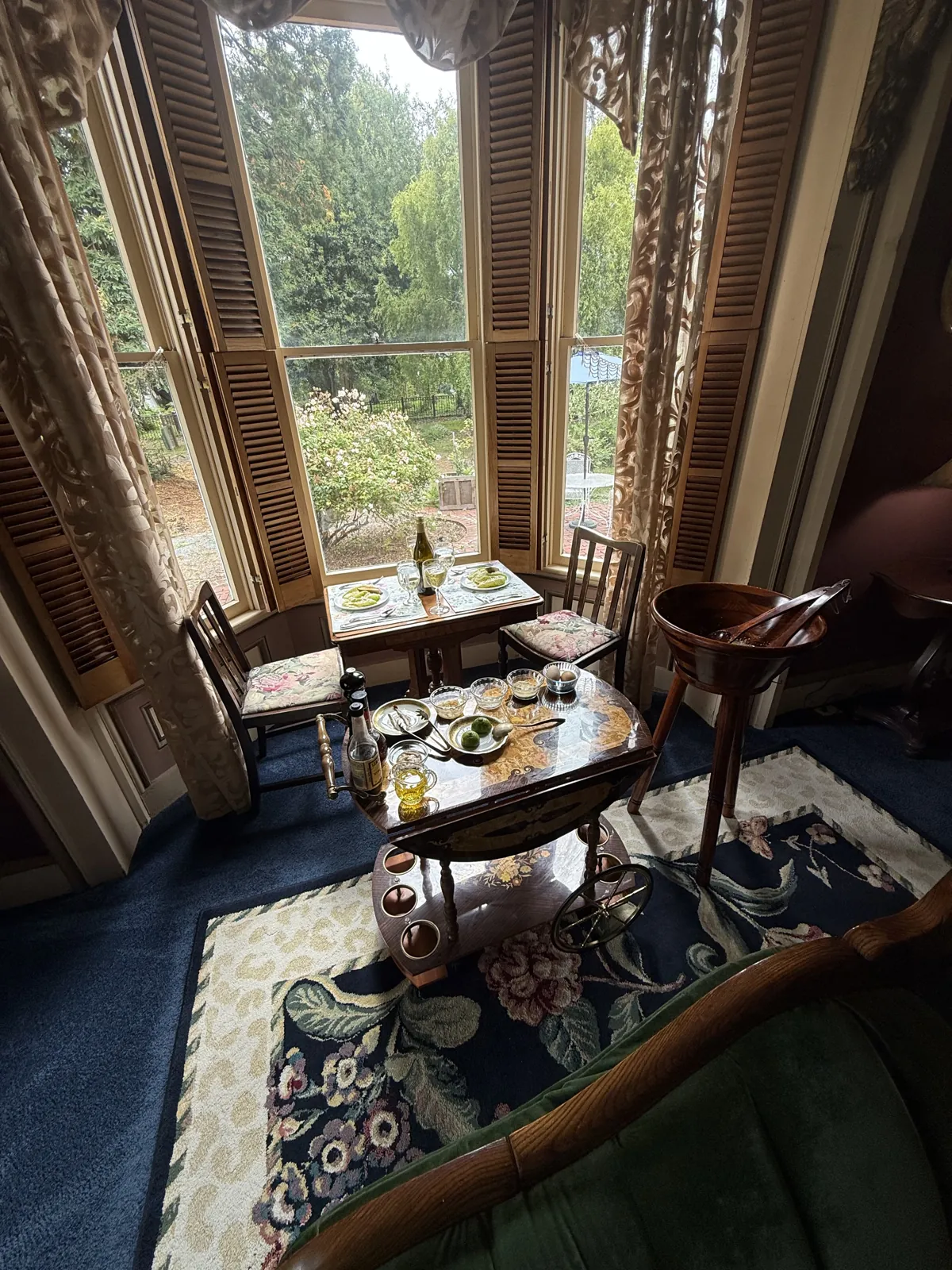 A table for two set with wine in a tall shuttered bay window overlooking the garden