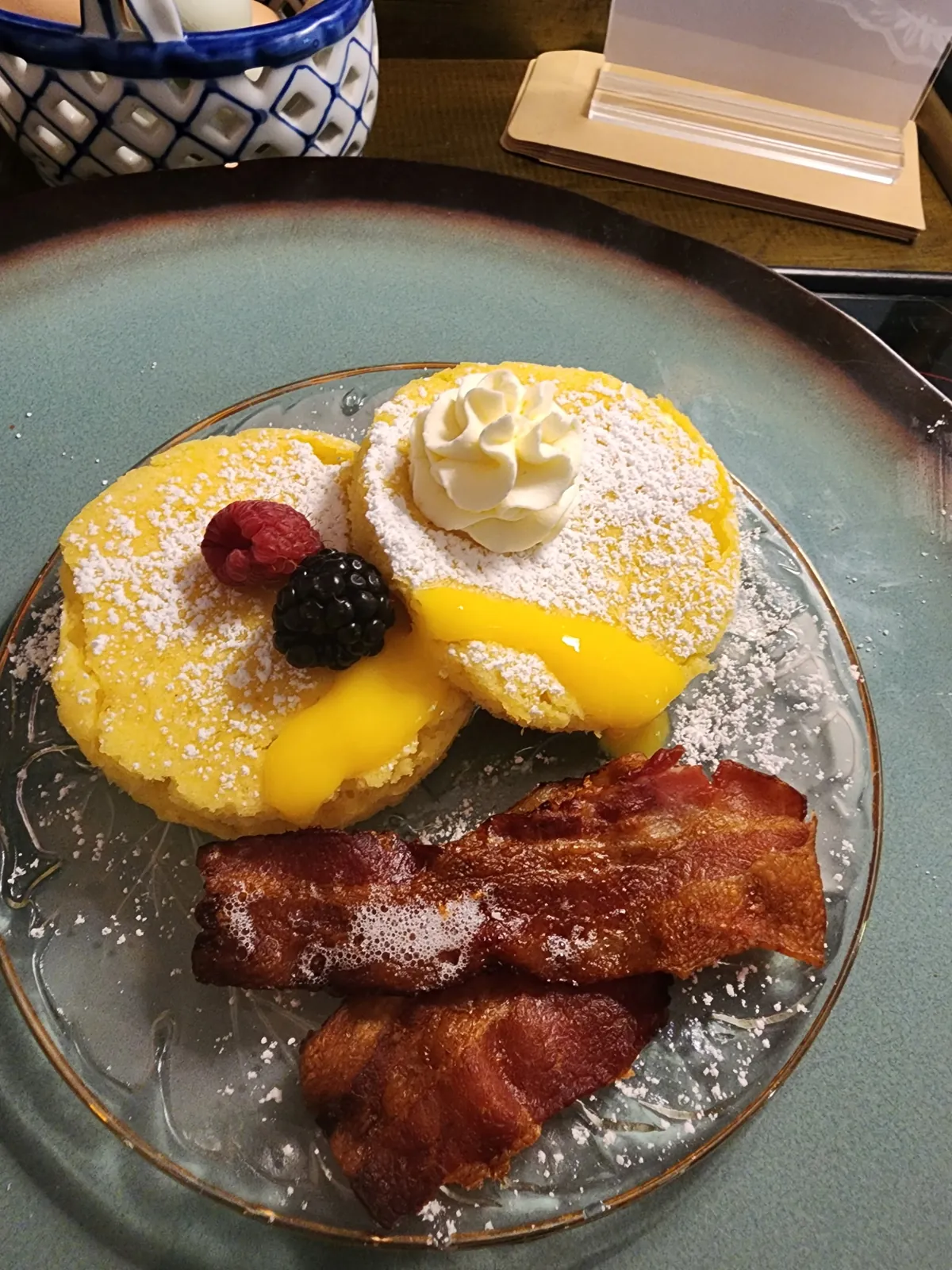 Breakfast plate of golden cakes with lemon curd, whipped cream, fresh berries, and bacon