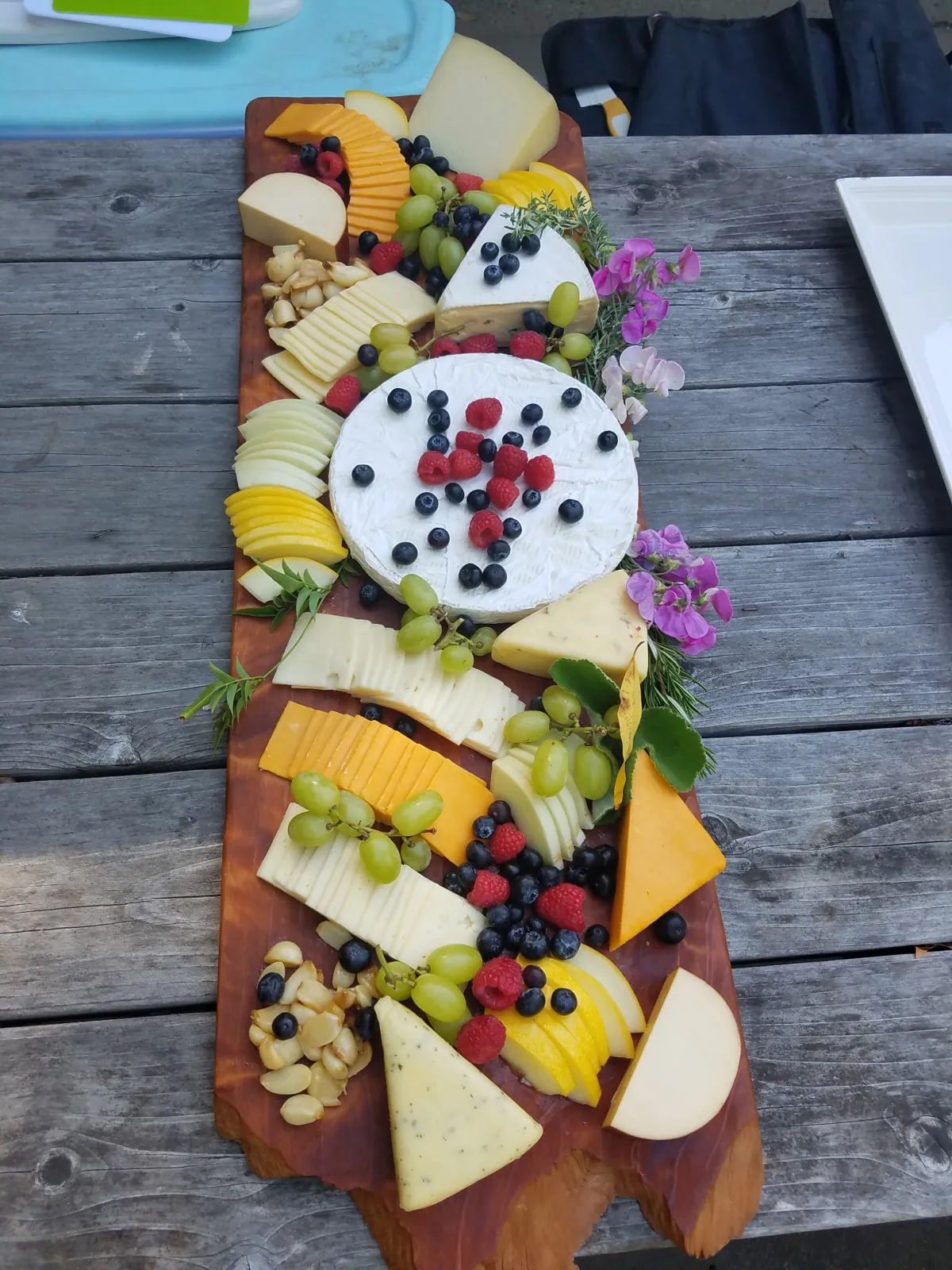 Long wooden board of assorted cheeses, grapes, berries, and flowers on an outdoor table
