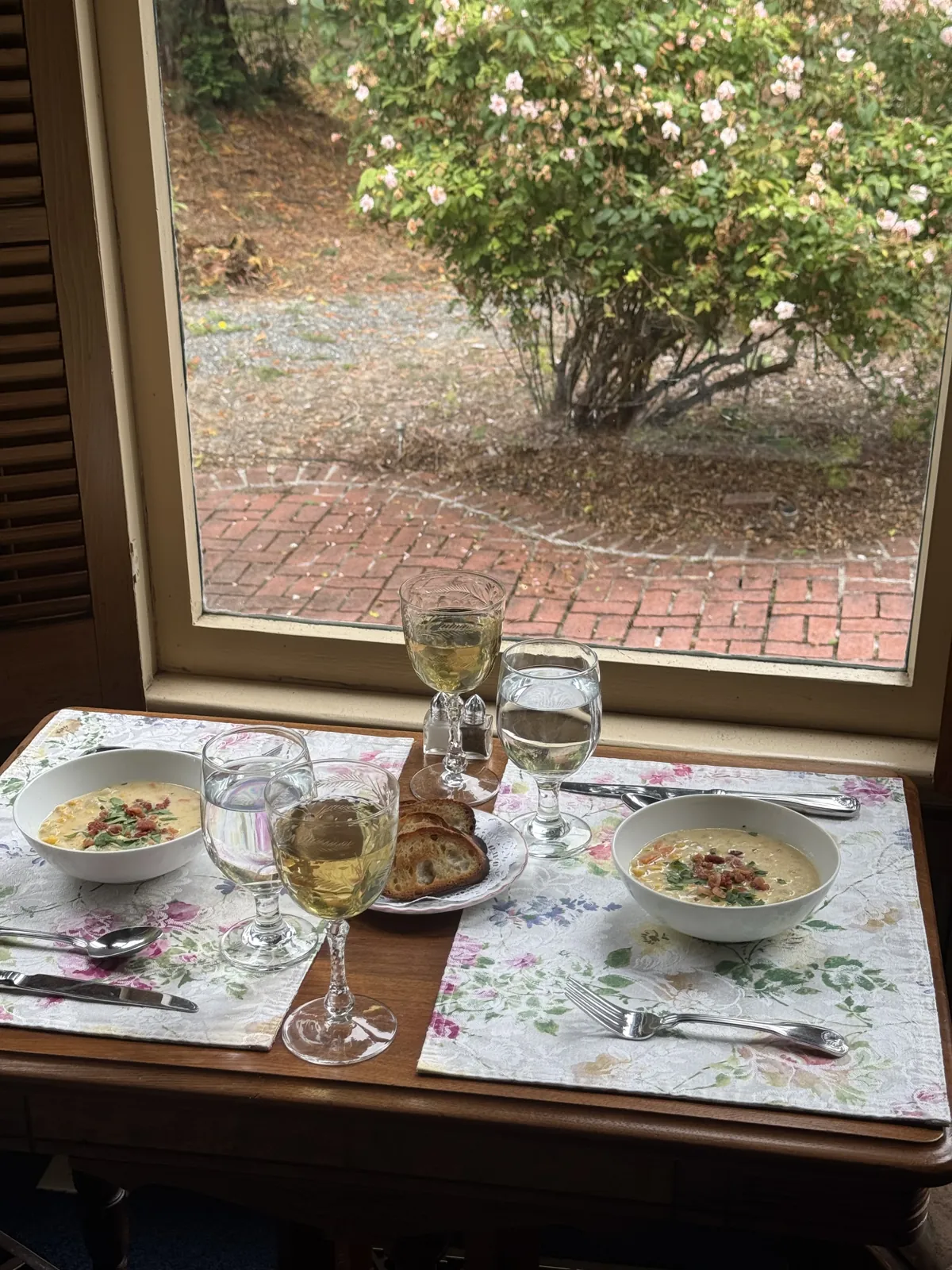 Two bowls of soup, toasted bread, and glasses of white wine on a table by a window facing the brick courtyard